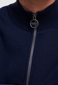 Námořnická zipová mikina s vysokým límcem, s kovovým zipem a kulatým poutkem na logu. Měkká textura s žebrovanými manžetami.