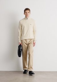 Light beige zip-up top paired with beige trousers. The model holds a black bag, wearing black shoes, against a minimalistic background.