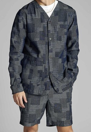 Mies pukeutunut siniseen ja harmaaseen teksturoidun patchwork-takin ja yhteen sopivien shortsien kanssa, seisoo yksi käsi taskussa ja toinen rentona.