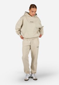 Μπεζ φούτερ με τσέπη καγκουρό και κέντημα. Συνδυάζεται με παντελόνια φόρμας που διαθέτουν ελαστική ζώνη και λογότυπα.