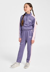 Lilla atletisk træningsdragt med en top med halv lynlås, pink side-striber og matchende løbebukser. Stoffet er glat og elastisk. Sølvfarvede atletiksko fuldender looket.