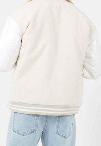 En person som bär en krämfärgad varsityjacka med vita ärmar och ljusblå jeans, sedd bakifrån.