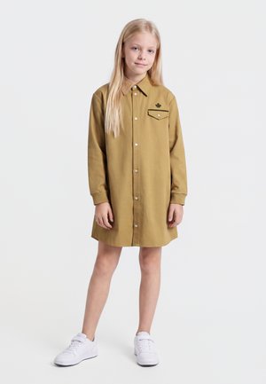 Unge pige med langt blondt hår iført en beige knap-op kjole med lomme detaljer og hvide sneakers, stående på en hvid baggrund.