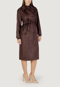 Bruine suède trenchcoat met een ceintuur in de taille, dubbele knopen, lange mouwen en een knielange snit. Draagt met naakte hoge hakken.