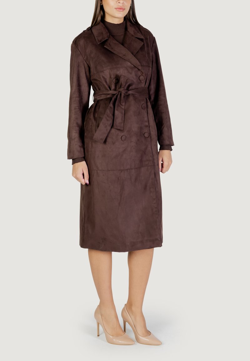 Bruine suède trenchcoat met een ceintuur in de taille, dubbele knopen, lange mouwen en een knielange snit. Draagt met naakte hoge hakken.