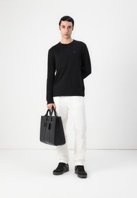 Schwarzes langärmliges Hemd, weiße Hose und schwarze Sneaker. Das Modell hält eine strukturierte schwarze Ledertasche mit zwei oberen Griffen und Akzenten.