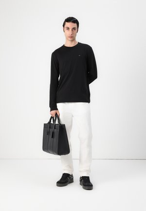Svart långärmad skjorta, vita byxor och svarta sneakers. Modellen håller en strukturerad svart läder tote med två handtag och detaljer.