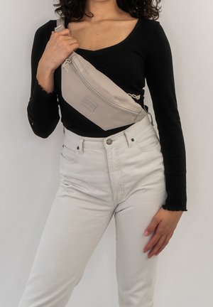 Personne portant un haut à manches longues noir et un pantalon clair, tenant un sac bandoulière beige devant sa poitrine.