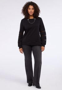 Mujer con cabello rizado que lleva un suéter negro con paneles de mangas transparentes, pantalones de campana oscuros y zapatos negros, de pie contra un fondo blanco.