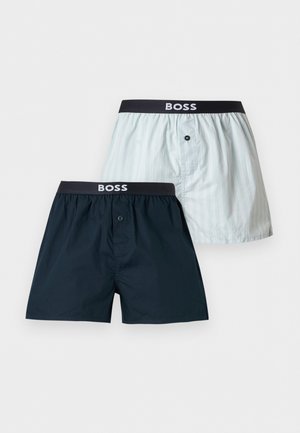 Ing nélküli férfi áll mezítláb fehér háttér előtt, sötétkék BOSS boxeralsót visel, egyik kezével igazítja a derékrészt.