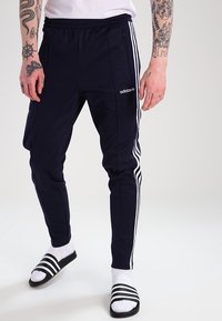 Person som bär marinblå Adidas träningsbyxor med vita ränder, vita strumpor och svarta sandaler. Hen visar upp tatuerade underarmar och händer.