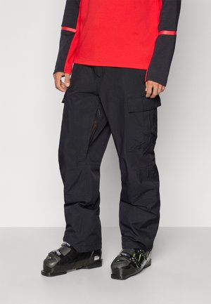 Pantalons cargo noirs avec plusieurs poches, fabriqués à partir d'un matériau durable. Portés avec des bottes de ski ; associés à une chemise à manches longues rouge et marine.