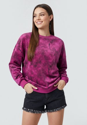 Smilende kvinde med langt brunt hår iført en lilla tie-dye sweatshirt og sorte slidte denimshorts, hænderne i lommerne.