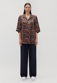 Camicia a maniche corte con stampa leopardata marrone e dettagli blu, con bottone frontale, abbinata a pantaloni larghi neri e sneakers bianche.