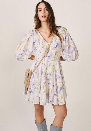 Mujer con vestido blanco de flores y mangas abullonadas, bolso cruzado beige y botas gris con tachuelas, mirando hacia adelante sobre un fondo liso.