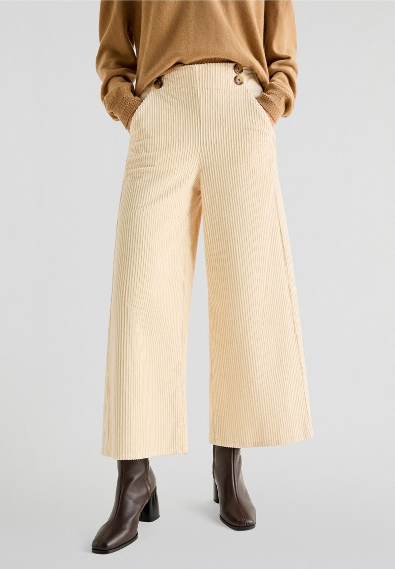 Personne portant un pantalon large beige en velours côtelé avec des détails de boutons et des bottines à talons marron foncé sur un fond uni.