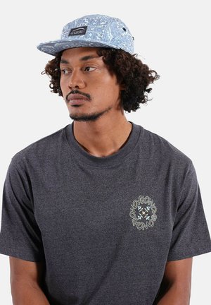 Homme aux cheveux bouclés portant une casquette bleue à motifs et un t-shirt gris foncé avec un graphisme circulaire sur le côté gauche de la poitrine, regardant vers la gauche.