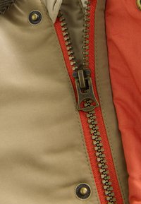 Gros plan d'une veste marron partiellement dézippée avec une doublure de fermeture éclair rouge et des boutons-pression en laiton visibles.