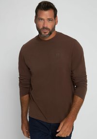 Camisa marrón de manga larga en un tejido suave con cuello redondo y un sutil logo en relieve en el pecho. Combinada con jeans oscuros.