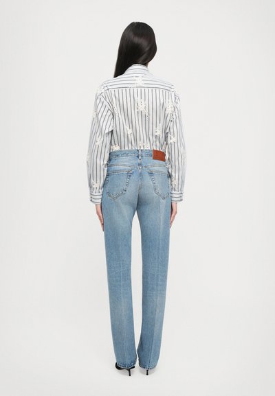 Črtasta srajca z dolgimi rokavi in cvetličnim vezenjem, v kombinaciji s svetlo modrimi hlačami z visokim pasom, ki imajo usnjeno oznako na hrbtu.