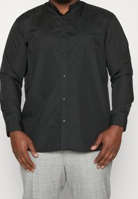 Zwarte overhemd met knopen en een mandarijnkraag, lange mouwen en een gladde textuur, gecombineerd met grijze geruite broek.