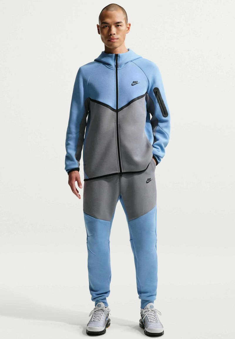 Tuta Nike blu e grigia con felpa con cappuccio e zip, design aderente, pannelli in tessuto a contrasto e tasca con zip sul braccio sinistro.