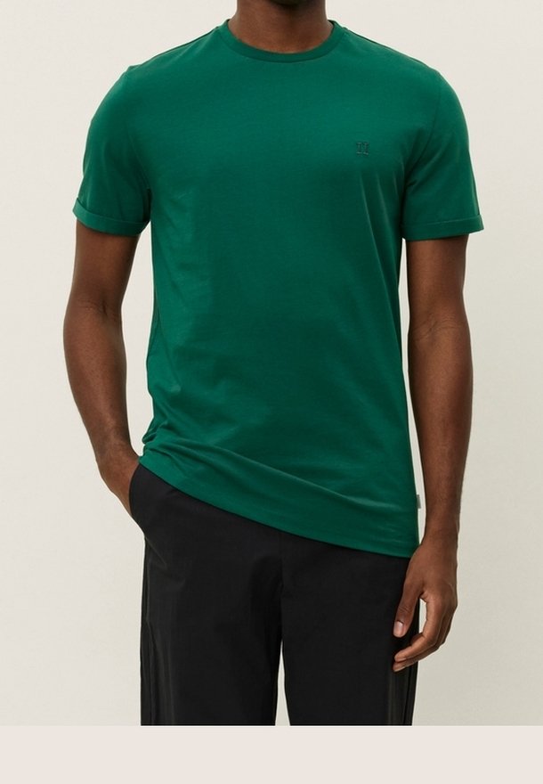 Homme portant un t-shirt vert à manches courtes avec un col rond et un pantalon noir, debout avec une main dans la poche et l'autre détendue.