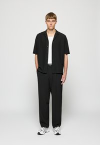 Jonge man staand met handen in zijn zakken, gekleed in een zwart shirt met korte mouwen, wit ondershirt, zwarte broek en witte sneakers tegen een effen achtergrond.