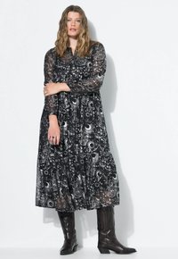 Donna con capelli mossi che indossa un lungo vestito nero con stampa celeste bianca e stivali neri a metà polpaccio, in piedi contro un muro bianco.
