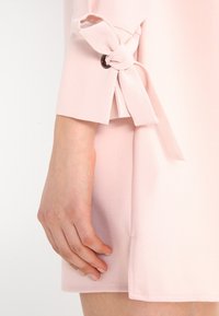 Różowa sukienka o gładkiej fakturze, z wiązaniem w supeł przy mankiecie i czarnym okrągłym akcentem. Krótki rękaw.
