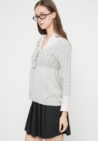 Νεαρή γυναίκα που φορά γυαλιά με καφέ-κεχριμπαρένιο σκελετό, ανοιχτό γκρι πουλόβερ πλεκτό με κόμπους πάνω από ανοιχτό ριγέ πουκάμισο και πλισέ σκούρα φούστα, σε λευκό φόντο.