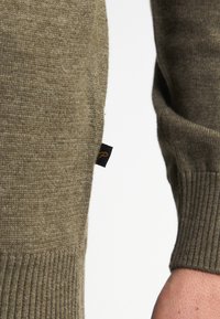 Olivgrüner Strickpullover mit geripptem Bündchen und einem schwarzen Etikett mit Logo an der Seite. Glatte Textur, schmaler Schnitt, sichtbare Nahtdetails.