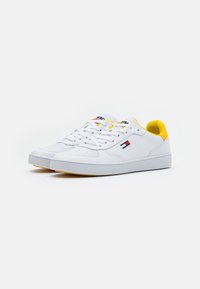 White sneakers with yellow accents, smooth leather upper, flat sole, and lace-up design. Features logo detailing on the side and tongue.