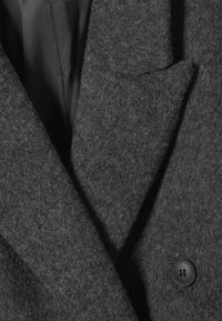 Mørk grå ullfrakk med et teksturert stoff, spiss krage og en enkelt svart knappdetalj foran. Polstringen er synlig under.