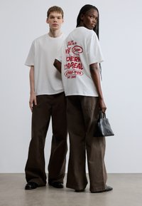 Camisetas blancas con texto gráfico rojo, combinadas con pantalones marrones de pierna ancha. Una persona lleva un pequeño bolso negro. Fondo neutro.