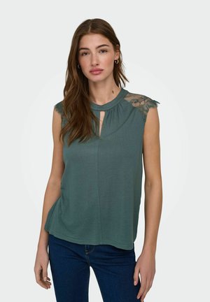 Kvinde med langt brunt hår iført grønt ærmeløst top med blonder på skuldrene og mørkeblå jeans, stående mod en ensfarvet lys baggrund.