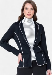 Crna blazer jakna s bijelim obrubom, slim fit kroj, dva prednja džepa, u paru s bijelim i crnim prugastim rolkom. Glatka tekstura tkanine.