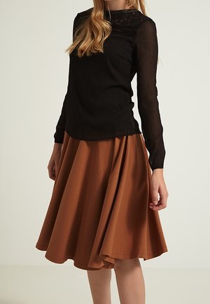 Femme portant un haut noir à manches longues en dentelle, associé à une jupe marron évasée longueur genou, debout devant un fond uni.