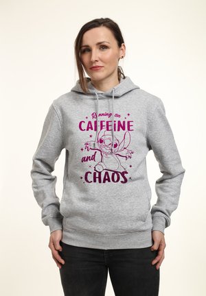 Donna che indossa una felpa grigia con cappuccio con testo viola e disegno animato che dice "Corro a caffeina e caos", in piedi su sfondo bianco.