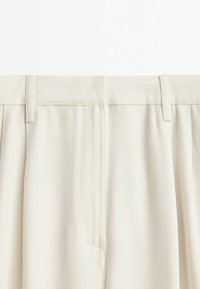 Gros plan sur la ceinture et la partie supérieure d'un pantalon crème avec passants pour ceinture et fermeture éclair dissimulée à l'avant.