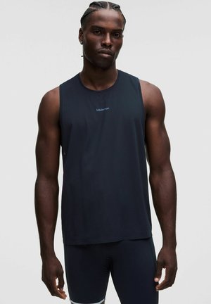 Muskuløs mand med flettet hår iført en sort ærmeløs Lululemon-trøje og sorte shorts, stående mod en ensfarvet baggrund.