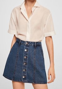 Camicetta corta beige chiaro abbinata a una gonna di jeans blu con chiusura frontale a bottoni, dettagli di cucitura e silhouette svasata.