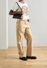 Béžové bavlněné kalhoty, krémové tričko s grafickým motivem motýla a černá kabelka přes rameno. Černé boty s masivní podrážkou. Přírodní dřevěná podlaha.