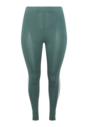 Person iført blågrønne leggings med hvide sidelommer, hvide sokker og hvide New Balance-sneakers, der står med den ene hånd hvilende på hoften.