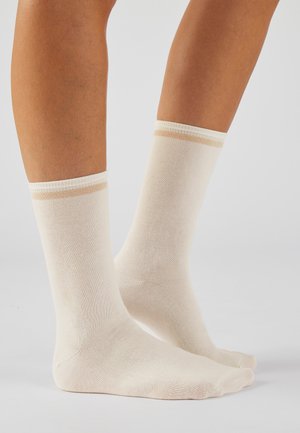 Gambe con calzini a crew color crema con una sottile striscia beige vicino alla parte superiore, in piedi su una superficie liscia grigio chiaro.