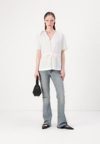Weißes Kurzarm-Bluse mit gebundenem Taillengürtel, kombiniert mit hellblauen, ausgestellten Jeans. Das Model hält eine schwarze geometrische Handtasche; der Hintergrund ist schlicht weiß.