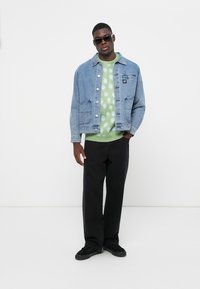 Denim jacket with two front pockets, light blue color; green sweater with white polka dots; black trousers; black sneakers; sunglasses.