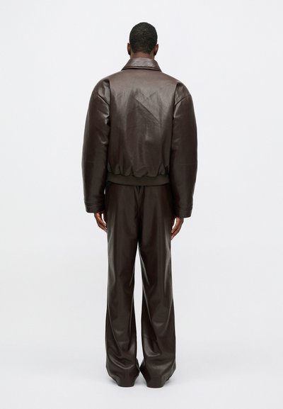 Jachetă din piele maro cu guler lat, manșete elastice și pantaloni evazați asortati, toate având o textură netedă și o croială lejeră.