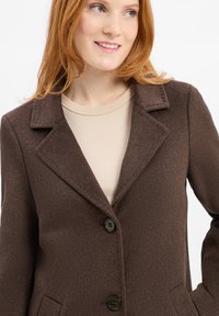 Cappotto in misto lana marrone scuro con colletto a rever, due bottoni, tasche frontali e finitura testurizzata. Indossato sopra un top beige.