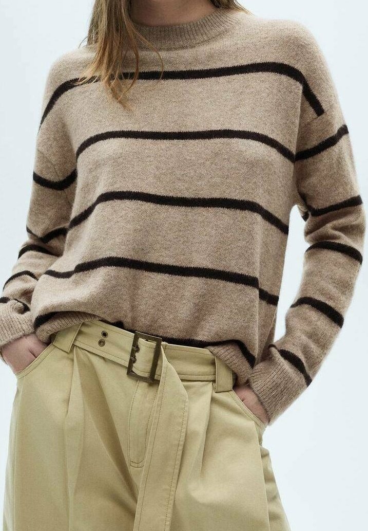 Femme portant un pull beige à rayures horizontales noires et un pantalon beige taille haute avec une large ceinture, mains dans les poches.